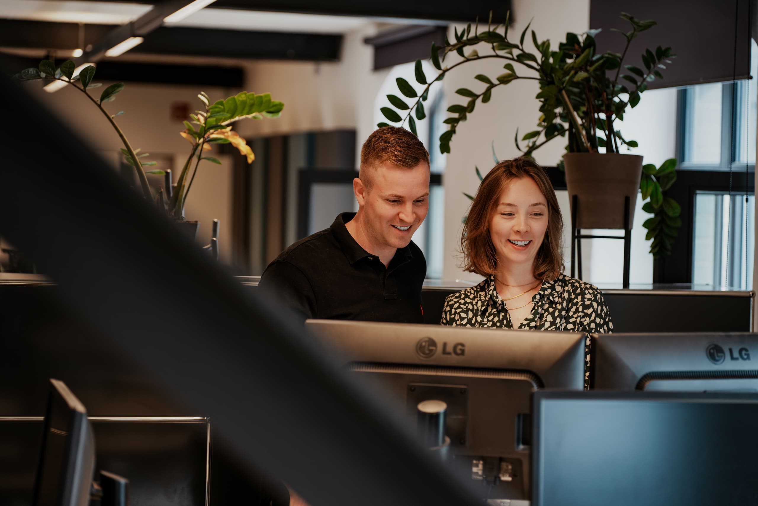 Zwei Mitarbeitende schauen lächelnd gemeinsam auf einen Monitor in einem modernen Büro